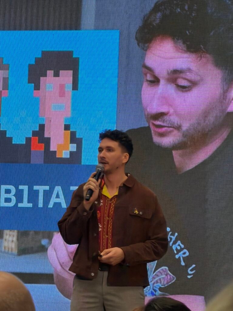 Pranešėjas scenoje konferencijoje „Šiauliai SMART 2025“, už jo ekrane pikselių stiliaus iliustracija